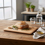 Loaf of bread on a wooden cutting board with a knife, surrounded by glasses and a carafe on a wooden table.