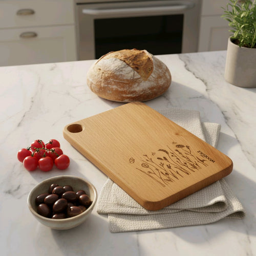 Wooden cutting board with engraved design on a kitchen counter with bread, tomatoes, and olives.