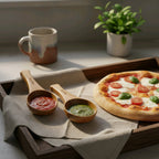 Wooden serving board with pizza, two wooden spoons with sauce, and a mug on a textured surface.